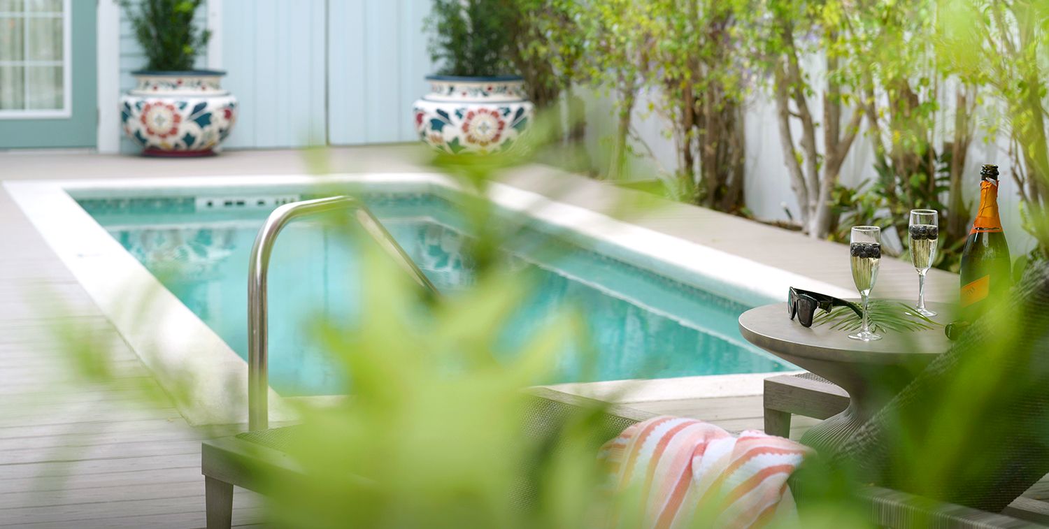 A quiet patio pool with greenery, patterned planters, and a table set with champagne and two glasses.