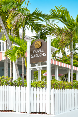 A &ldquo;DUVAL GARDENS&rdquo; entrance sign rises above tropical palm trees beside a white resort building and garden fence under a blue sky.