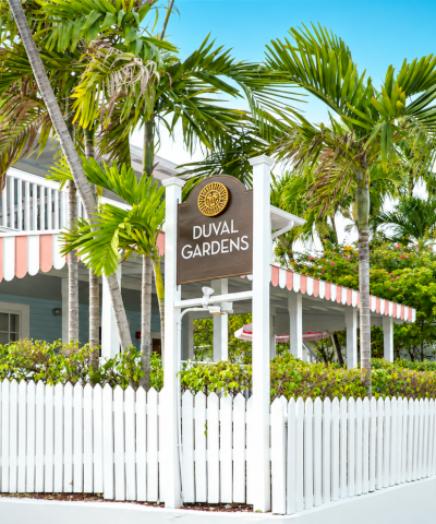 A &ldquo;DUVAL GARDENS&rdquo; entrance sign rises above tropical palm trees beside a white resort building and garden fence under a blue sky.