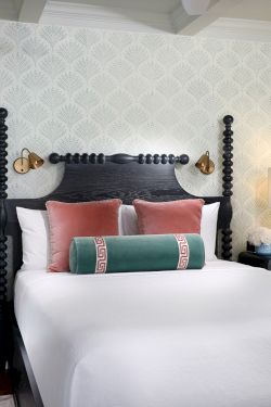 A stylish bedroom with a dark four-poster canopy bed, colorful pillows, bedside lamps, and patterned wallpaper, all in soft daylight.