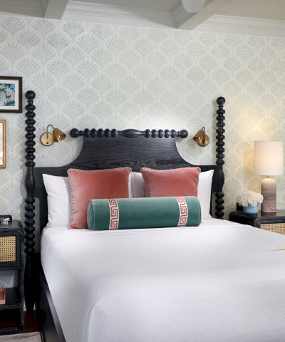 A stylish bedroom with a dark four-poster canopy bed, colorful pillows, bedside lamps, and patterned wallpaper, all in soft daylight.