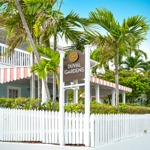 A sunny tropical entrance with palm trees and a sign reading &ldquo;DUVAL GARDENS&rdquo; by a white building and walkway lamp.