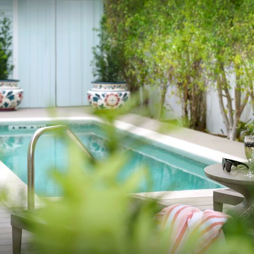 A poolside patio with champagne glasses and a bottle on a table, surrounded by greenery and lounge chairs near the pool.