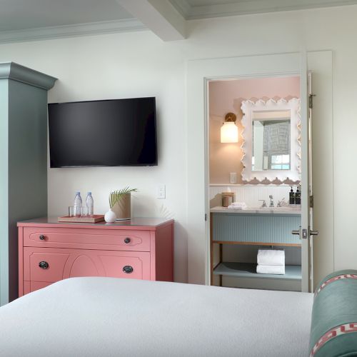 A bright bedroom with a mounted flat-screen TV, a pink dresser, and an open door to a tidy bathroom vanity and mirror.