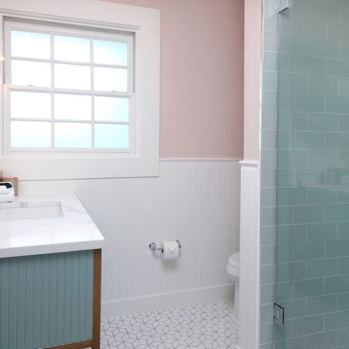 A bright bathroom with a window over a white sink, pastel walls, blue tiled shower, and chrome fixtures.