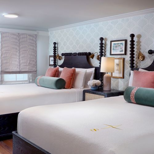 A bright modern bedroom with two made beds, dark carved headboards, patterned wallpaper, lamps, and decorative pillows.