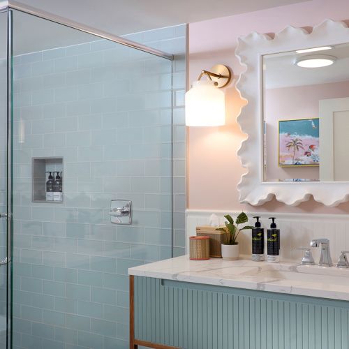 A modern bathroom with a glass shower, blue tiles, and a marble vanity lit by a scalloped mirror and wall sconce.