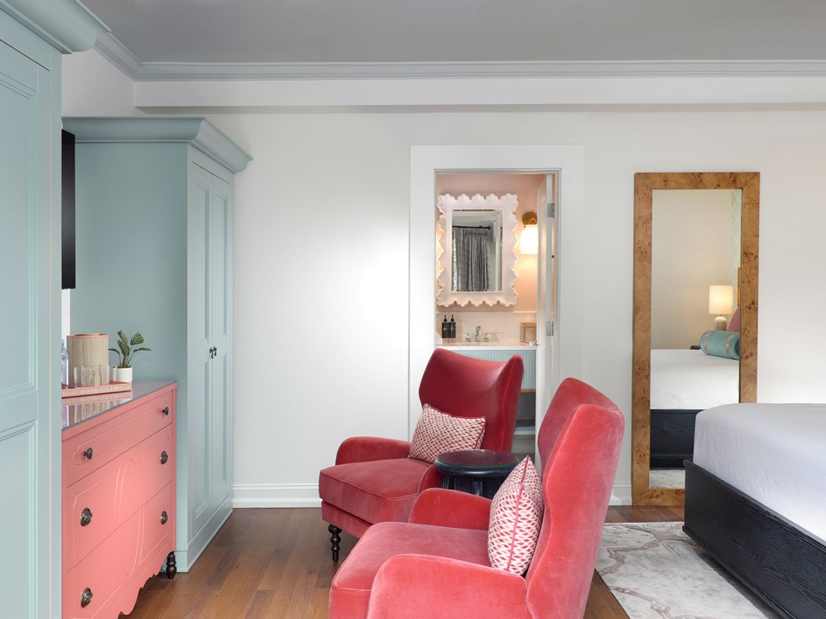 A bright modern room with pastel walls, a pink dresser, red armchairs, and a mirror revealing a small vanity area.