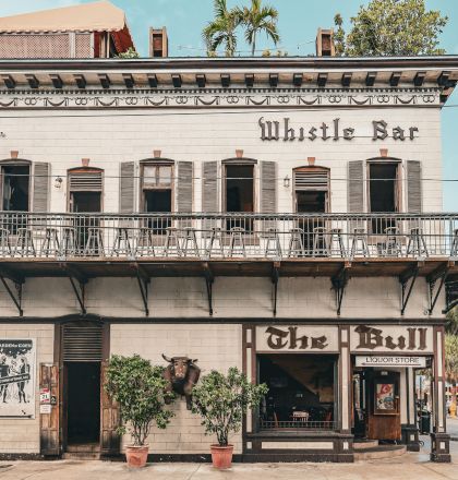 A weathered two-story building facade with ornate balcony trim, tropical plants, and sign text &ldquo;Whole Ban&hellip;&rdquo; and &ldquo;Liquor Store.&rdquo;