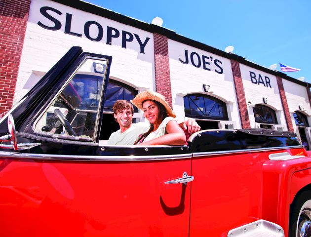 A couple rides in a red convertible outside Sloppy Joe&rsquo;s bar, with the &ldquo;Joe&rsquo;s&rdquo; sign visible behind them.