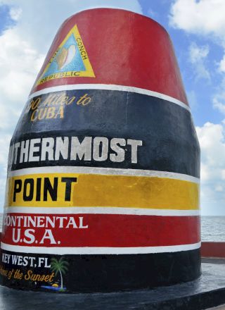 A painted buoys marked &ldquo;CUBA&rdquo; and &ldquo;OTHERMOST POINT CONTINENTAL U.S.A.&rdquo; stands by a spiral fence, with ocean and blue sky behind.