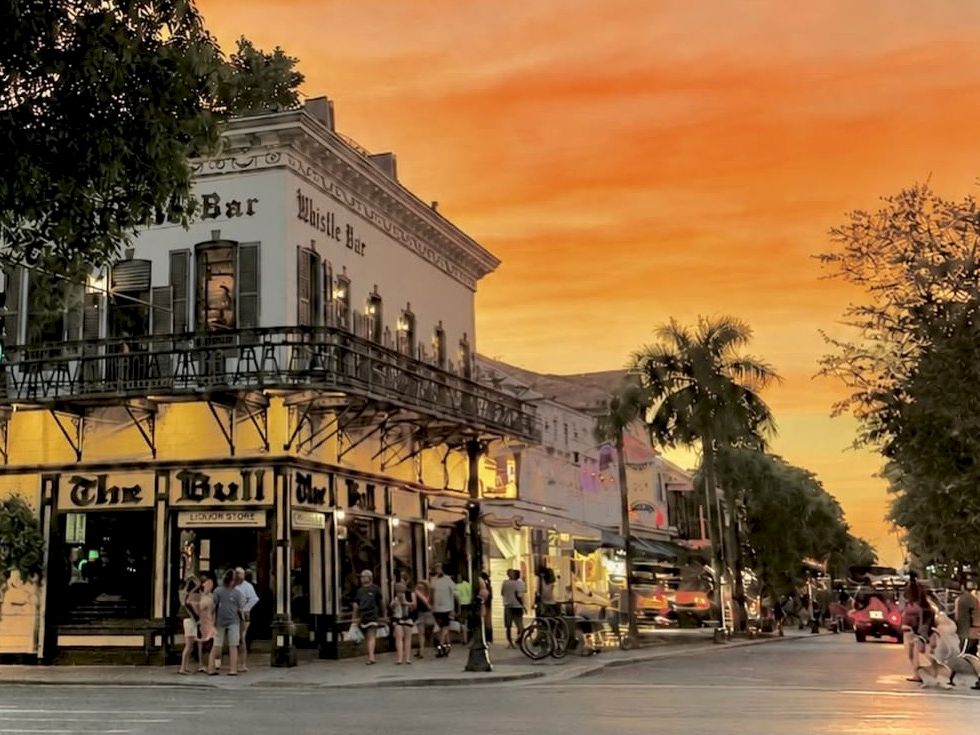 A street at sunset with the &ldquo;Bull and Bush&rdquo; bar storefront, people walking, palm trees, and cars parked.