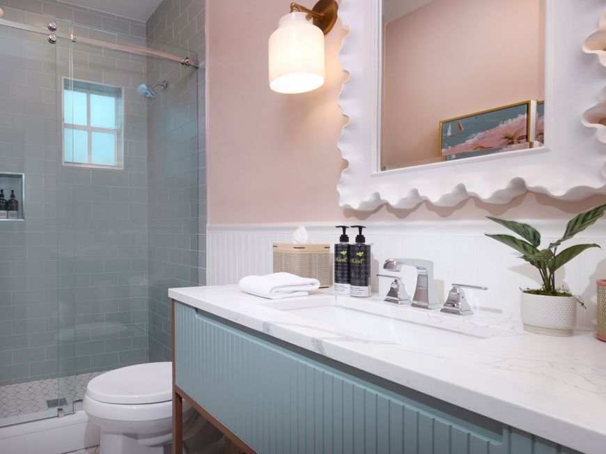 Bright modern bathroom with marble vanity, wavy mirror, toiletries, and a tiled walk-in shower.