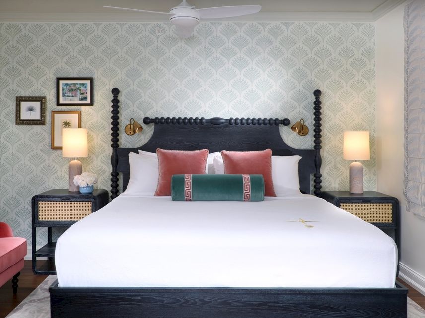 A well-styled bedroom with a dark upholstered bed, white linens, red chairs, bedside lamps, and patterned wallpaper.