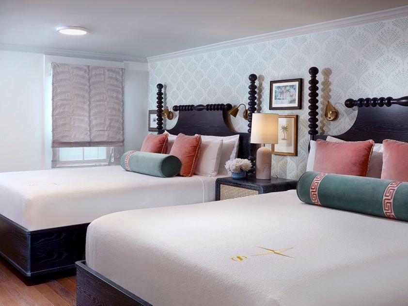 A bright bedroom with two beds, patterned wallpaper, orange pillows, green cushions, and bedside lamps.