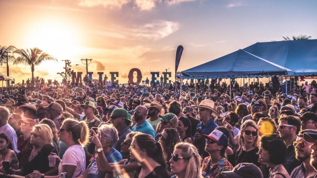 A packed outdoor music festival at sunset, with palm trees, big blue tents, and a cheering crowd filling the field in golden light.