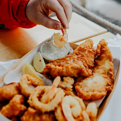 A hand dips a fried shrimp/chicken piece into a cup of creamy sauce while enjoying takeout by the seaside.