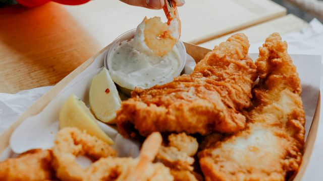 A hand dips a fried shrimp/chicken piece into a cup of creamy sauce while enjoying takeout by the seaside.