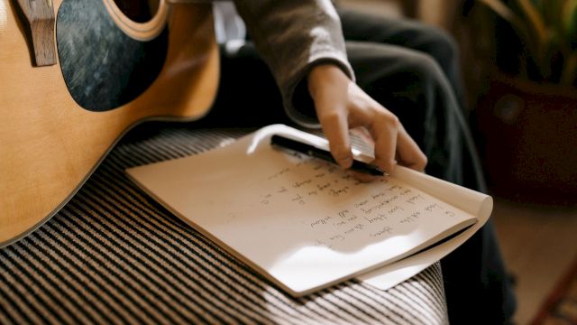 A person with an acoustic guitar scribbles notes on sheet music while holding a pen.