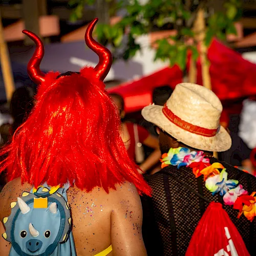 A person in a red devil outfit with bright red horns and wig at a lively outdoor festival, standing among other costumed revelers.