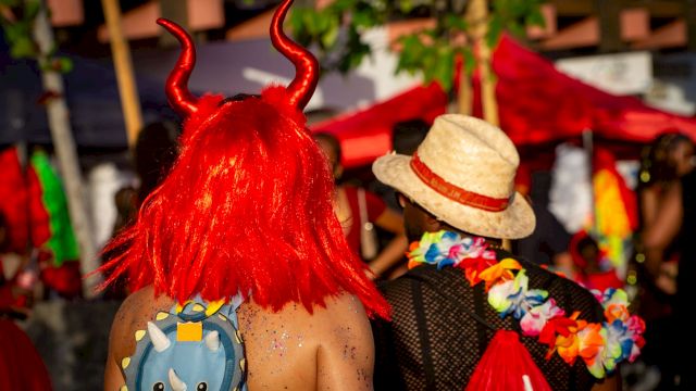 A person in a red devil outfit with bright red horns and wig at a lively outdoor festival, standing among other costumed revelers.