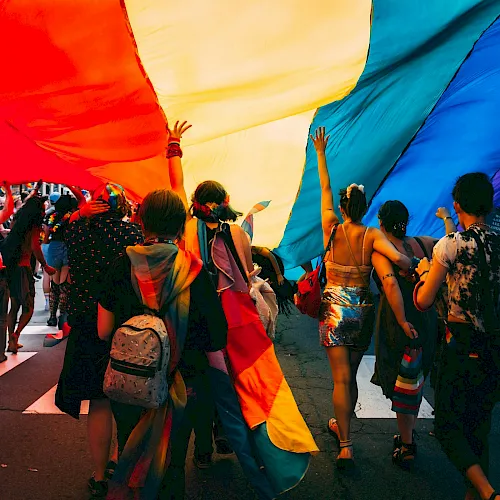 A lively crowd dances under bright red and blue festival tents, with people holding up their hands and celebrating together.