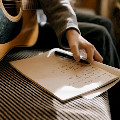 A person in a cozy room writes notes on paper with a pen while a guitar sits nearby.