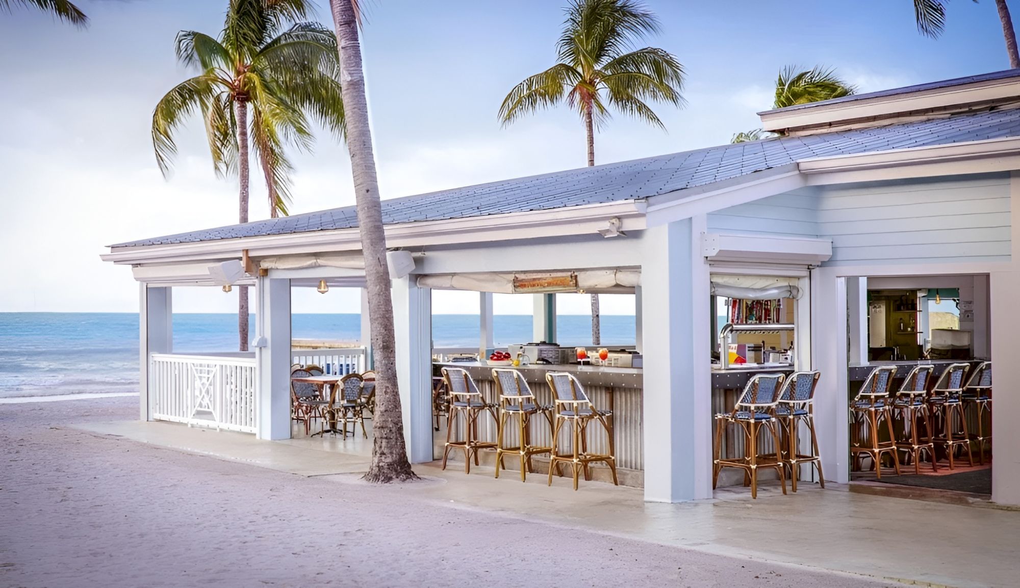 A beachside bar with gold chairs and stools, palm trees, and a view of the ocean under a clear sky.
