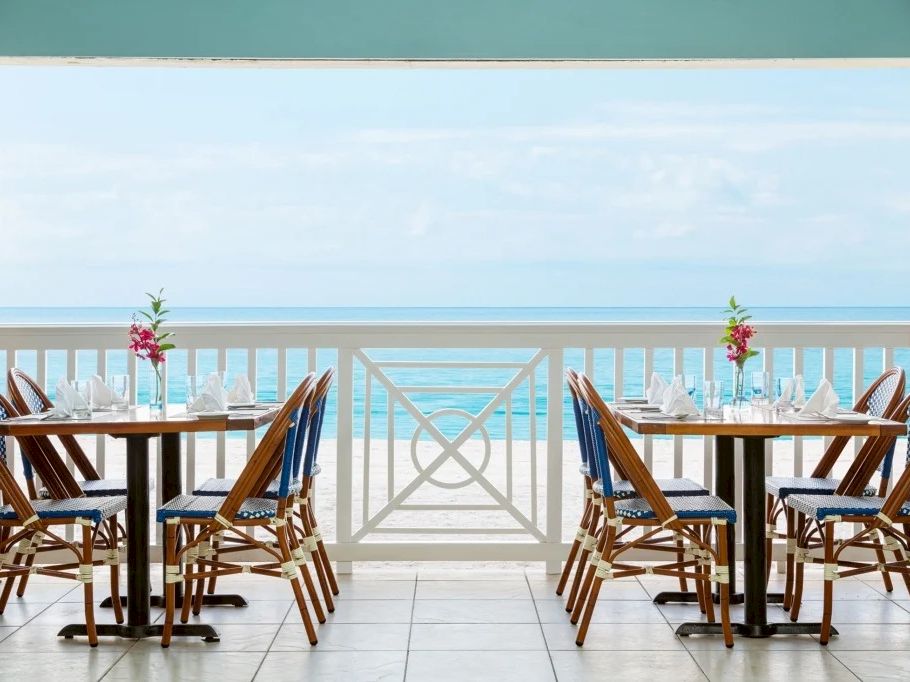 Beachfront patio dining set with two wooden tables and blue chairs, decorated with flowers, overlooking the ocean.
