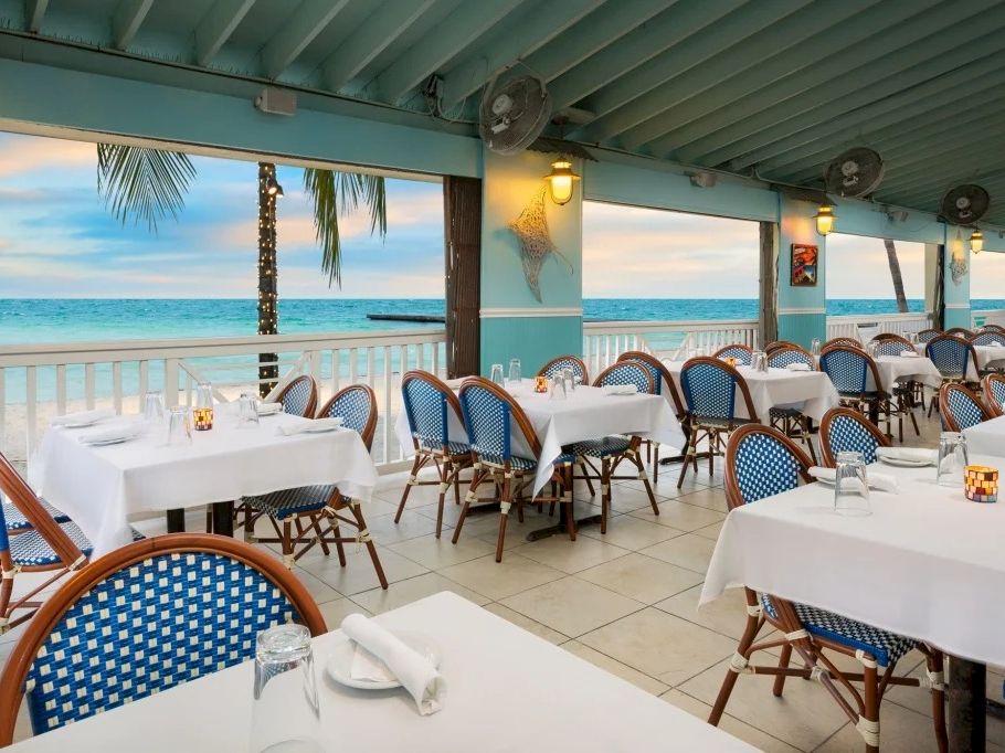 A seaside dining terrace with neatly set white tables and blue woven chairs, open to the ocean view through large windows.