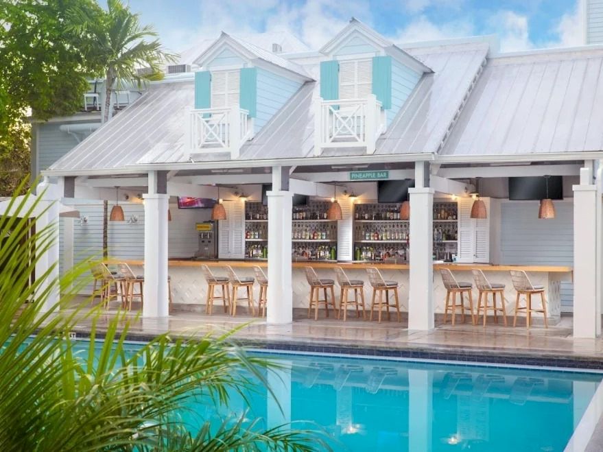 An outdoor poolside bar with white shutters, pendant lights, and palm plants under a sunny blue sky.