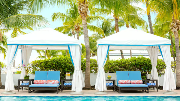 A tropical poolside scene with two white-and-blue cabanas, lounge chairs, and tall palm trees around a sparkling swimming pool.