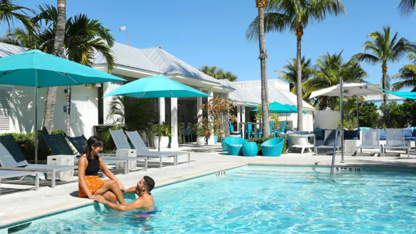 A sunny hotel pool with two people relaxing in the water, framed by palm trees, loungers, and turquoise umbrellas.